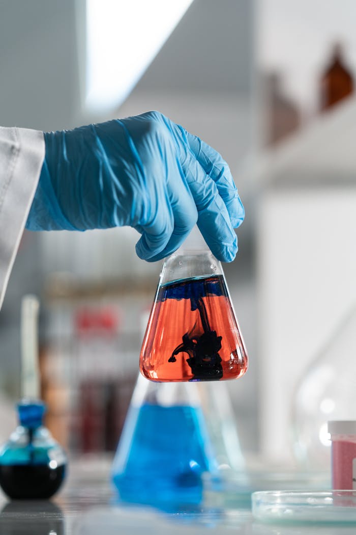 Close-up of hand in glove holding a flask with colored liquid in a laboratory setting.